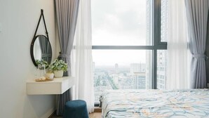 Quarto com uma janela ampla, mesa de cabeceira com espelho, uma pequena planta e um banquinho azul.