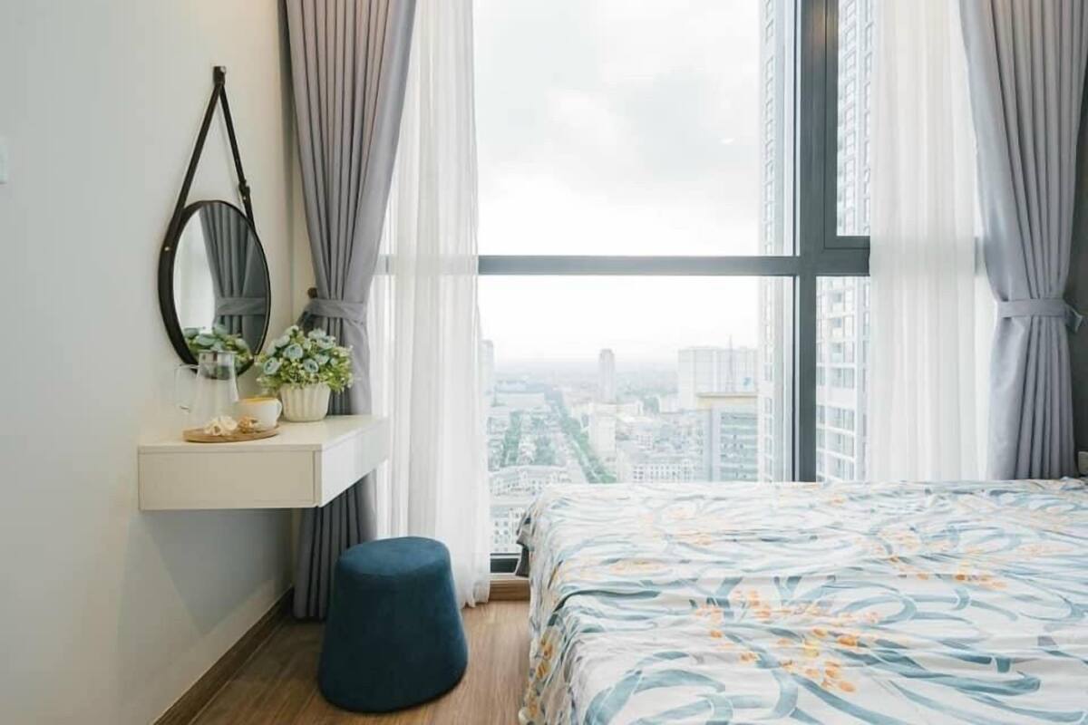 A bedroom with a large window, a bedside table with a mirror, a small plant, and a blue ottoman.