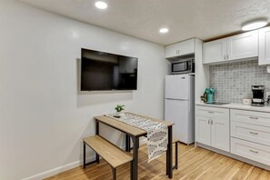 Une cuisine compacte avec des meubles blancs, un four à micro-ondes, un réfrigérateur, une table en bois avec un banc, et un téléviseur à écran plat fixé au mur.