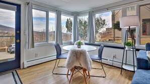 A room with a round table, two chairs, a white furry chair, a floor lamp, and large windows with a view of trees and a house.