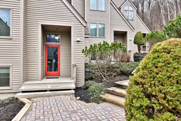 A two-story residential building with a red door, a stone wall, and a small tree in the front yard.