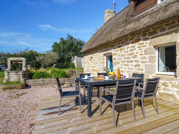 A stone cottage with a thatched roof, a wooden deck, and a table set for dining.