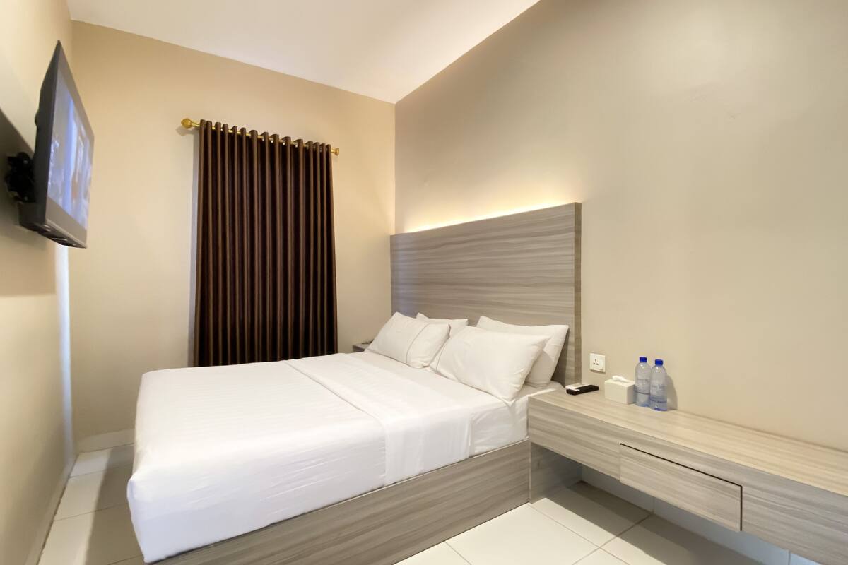 A modern hotel room with a large bed, a bedside table, a flat-screen TV mounted on the wall, and a window with curtains.