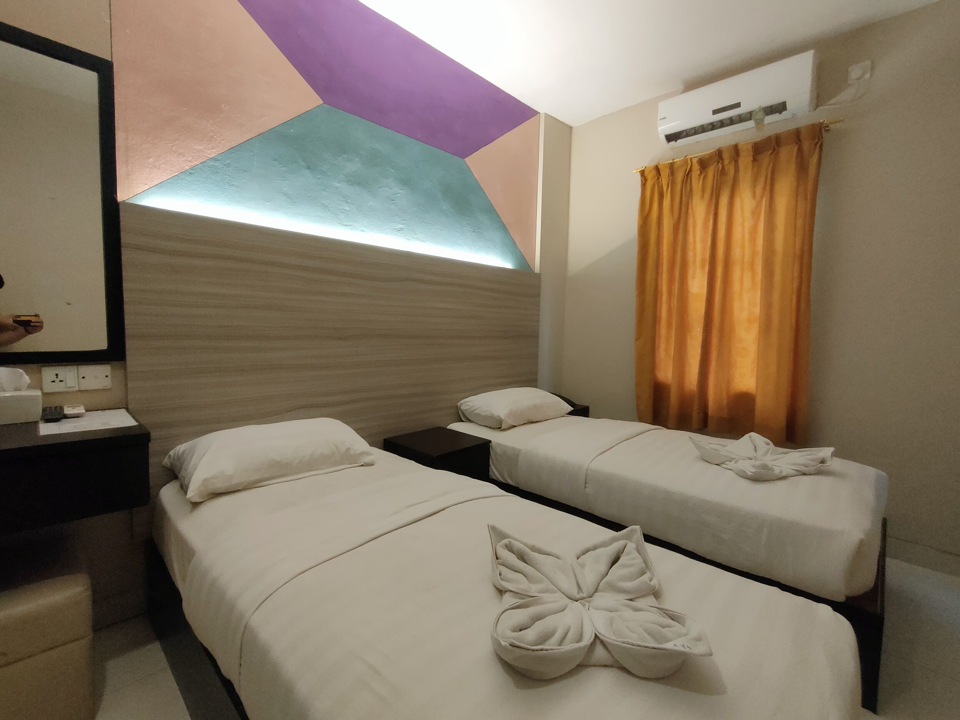 A hotel room with two single beds, a nightstand, a mirror, and an air conditioning unit.