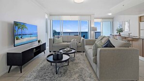 A modern living room with a large flat-screen TV, two sofas, a coffee table, and a view of the ocean through large windows. - Chevron Renaissance - WOWSTAY (Surfers Paradise)