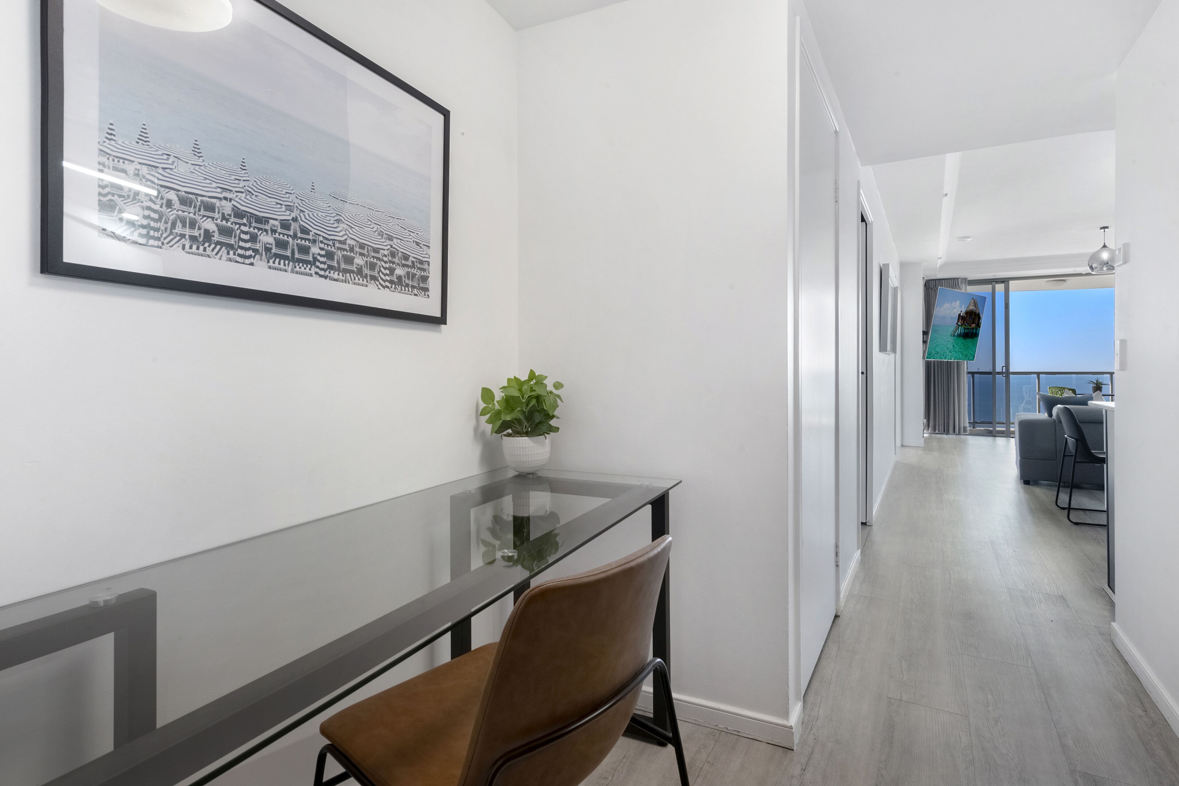 A modern hotel room with a glass table, a brown chair, a framed picture on the wall, and a view of the balcony.