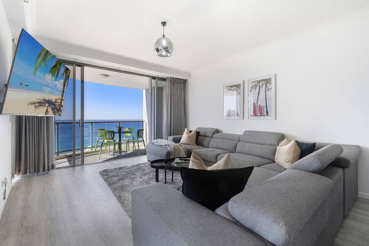 Un salon moderne avec un canapé d’angle gris, une table basse et un grand écran de télévision diffusant une scène de plage. Il y a un balcon avec vue sur la mer et une chaise à l’extérieur.
