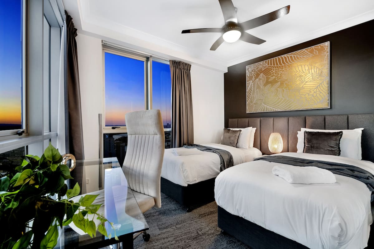 Une chambre d’hôtel avec deux lits, un ventilateur de plafond, une grande fenêtre avec des rideaux, une œuvre d’art accrochée au mur et une plante.