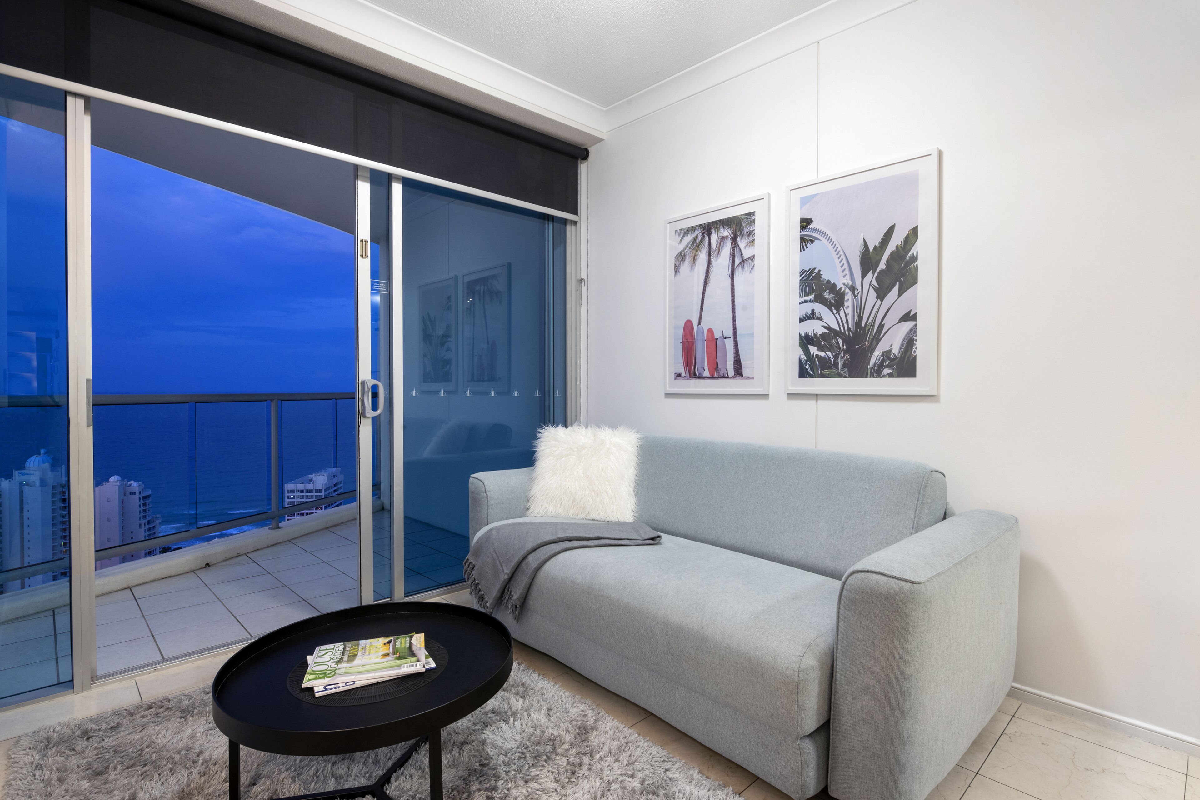 Un salon moderne avec un canapé gris, une petite table noire sur laquelle sont posés des magazines, un tapis gris moelleux et une grande porte-fenêtre coulissante donnant sur un balcon avec vue sur la ville.