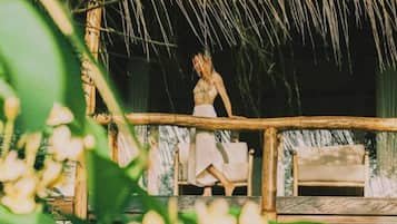 Una persona caminando sobre una terraza de madera con techo de paja, una silla y una mesa.