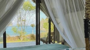 Een slaapkamer met een bed, gordijnen en uitzicht op zee en bomen.