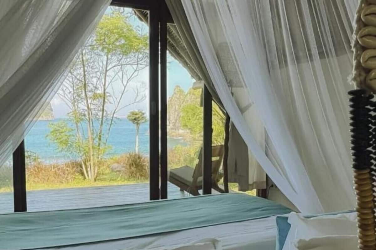 Een slaapkamer met een bed, gordijnen en uitzicht op zee en bomen.