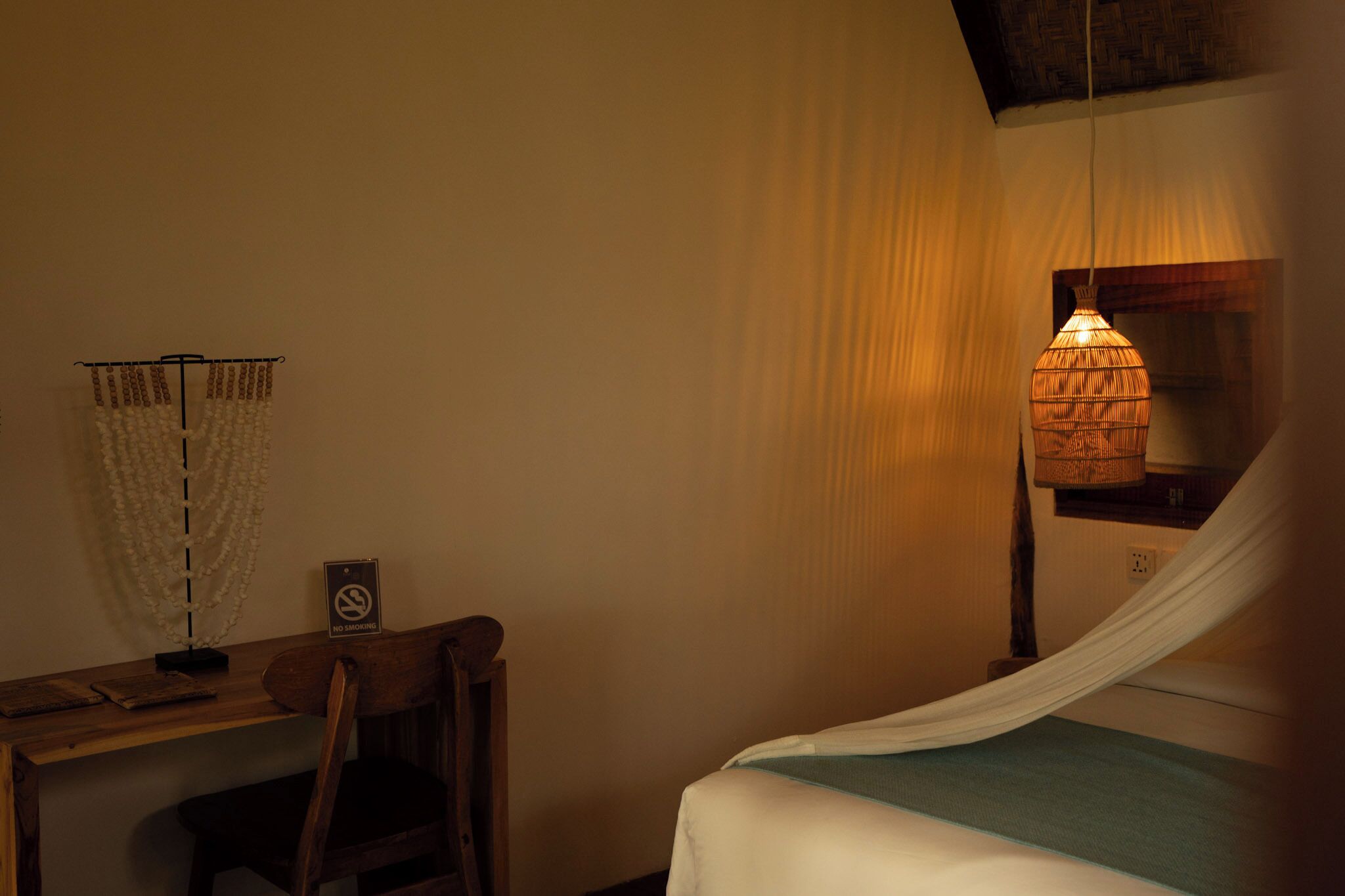 A room with a bed, a desk, a chair, a hanging light fixture, and a 'No Smoking' sign.