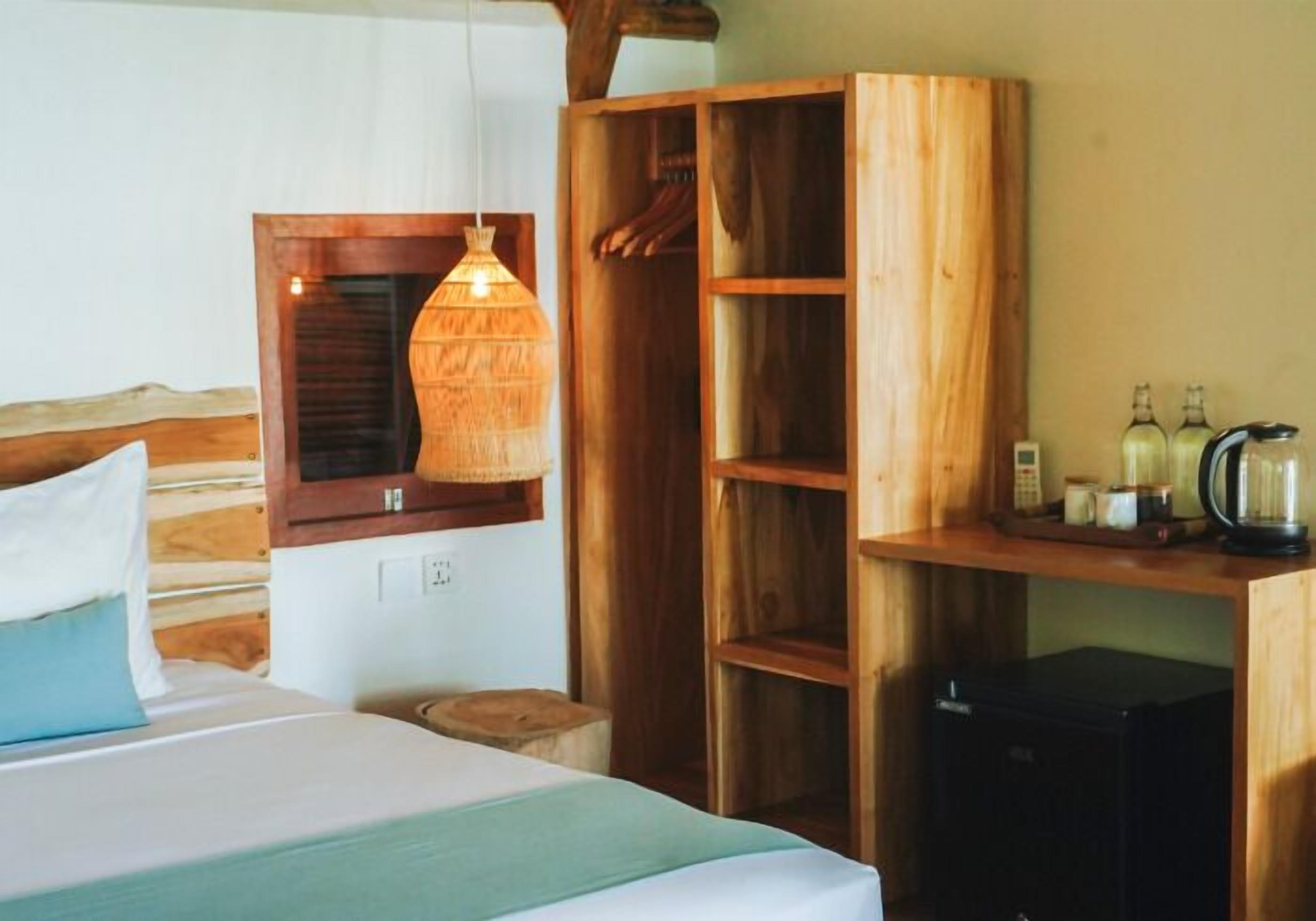 A bedroom with a bed, a wooden wardrobe, a hanging light fixture, and a coffee maker on a shelf.