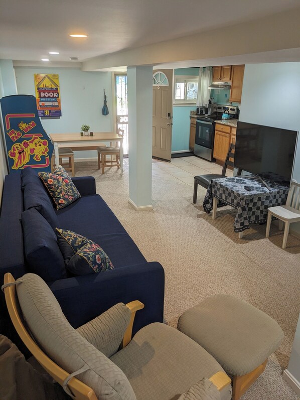 A living room with a blue sofa, a wooden chair, and a small table with a checkered tablecloth. There is a kitchen area with wooden cabinets and a stove.