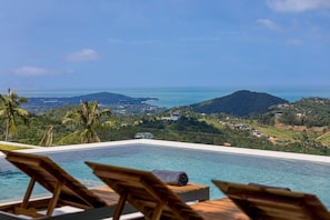 A pool with wooden loungers and a view of a coastal landscape with hills and a body of water. - Lilly Belle (Koh Samui)
