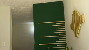 Ein modernes Zimmer mit einem Holztisch, zwei weißen Hocker, einer grünen Akzentwand und einem Bett im Hintergrund.
