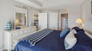 Une chambre à coucher comprenant un lit, une commode, un miroir, un ventilateur de plafond et une porte donnant accès à une salle de bain.