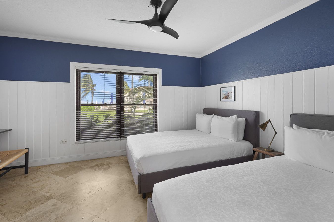A bedroom with two beds, a ceiling fan, a window with blinds, and a small table.