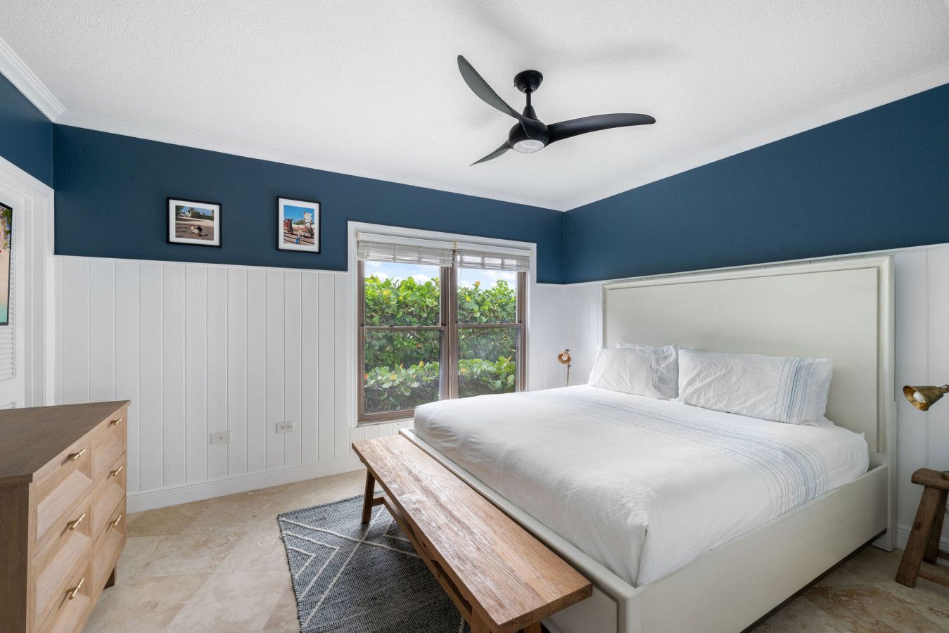 A bedroom with a bed, a ceiling fan, a window with a view of greenery, and a wooden dresser.