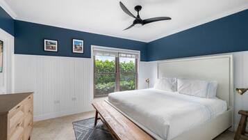 A bedroom with a bed, a ceiling fan, a window with a view of greenery, and a wooden dresser.