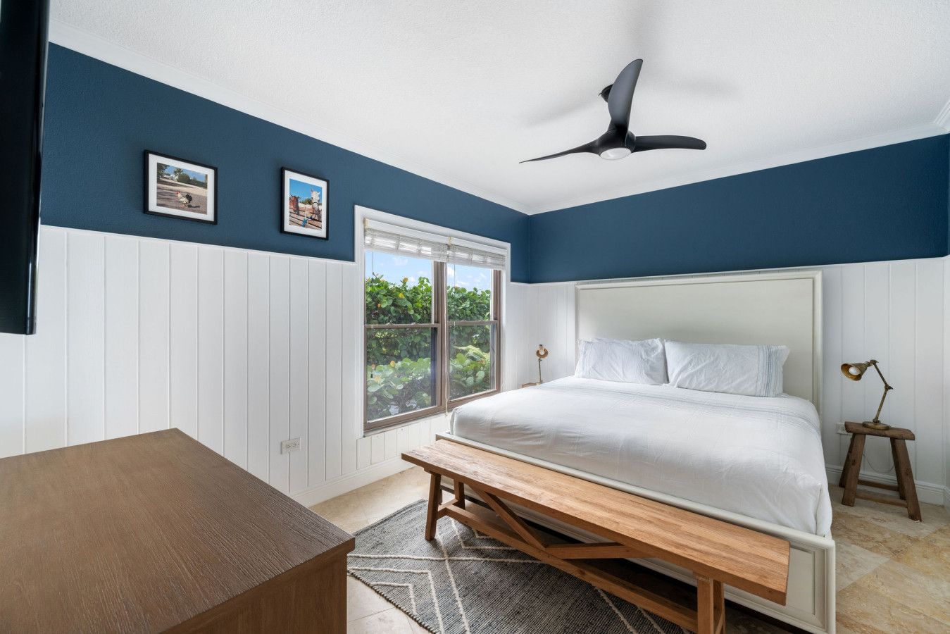 Quarto com cama, escrivaninha, ventilador de teto e janela com vista para o verde.