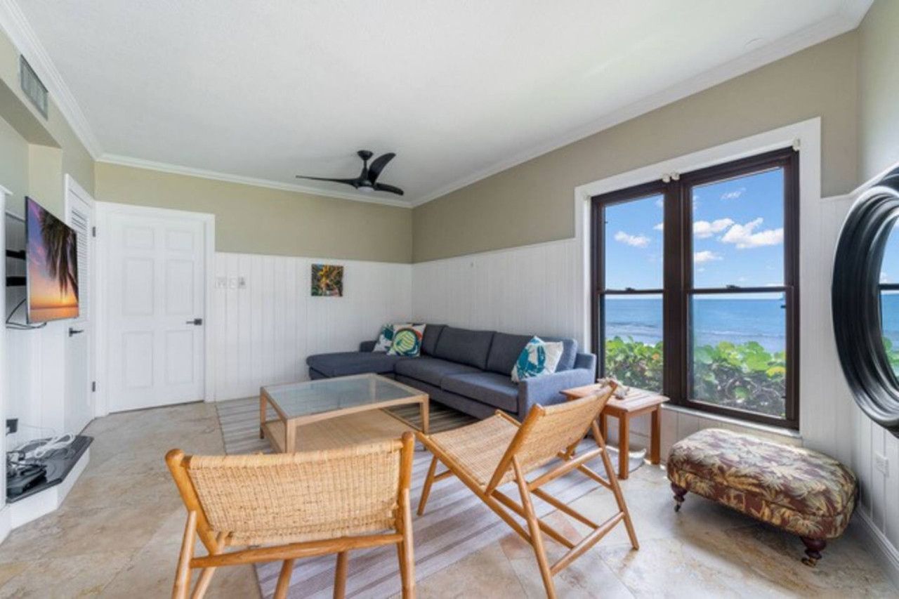 A modern living room with a sofa, a glass coffee table, wicker chairs, a ceiling fan, and a large window with a view of the ocean.