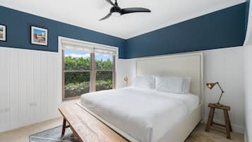 Un dormitorio con cama, ventilador de techo, ventana con vistas a la vegetación y un banco de madera.