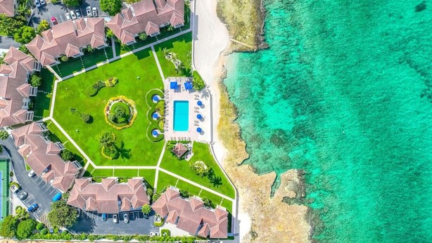 Une vue aérienne d’un complexe hôtelier avec une piscine centrale, entourée de pelouses verdoyantes et de palmiers, et donnant sur une plage aux eaux turquoise et cristallines.