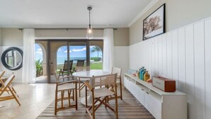 Un coin repas avec une table ronde, des chaises, un luminaire suspendu et une vue sur l’océan par une porte vitrée.