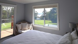 Un dormitorio con una cama grande, una silla de mimbre con cojines, una ventana que ofrece vistas a un área verde y una caravana, y una lámpara sobre una mesita de noche.