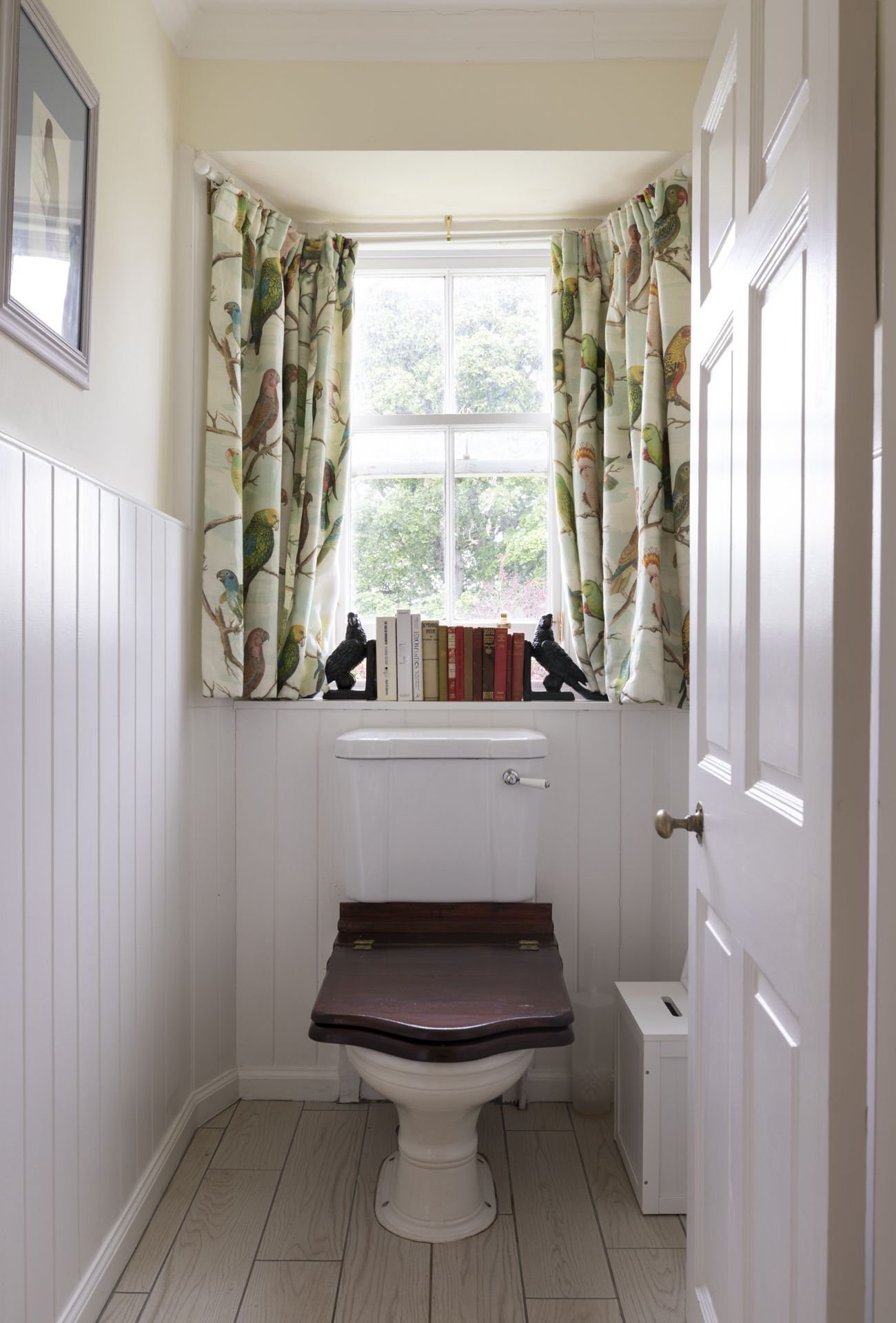 A small bathroom with a toilet, a window with curtains, and a shelf with books.