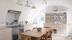 A kitchen with a stove, oven, and a dining table with chairs. There is a shelf with dishes and a clock on the wall.