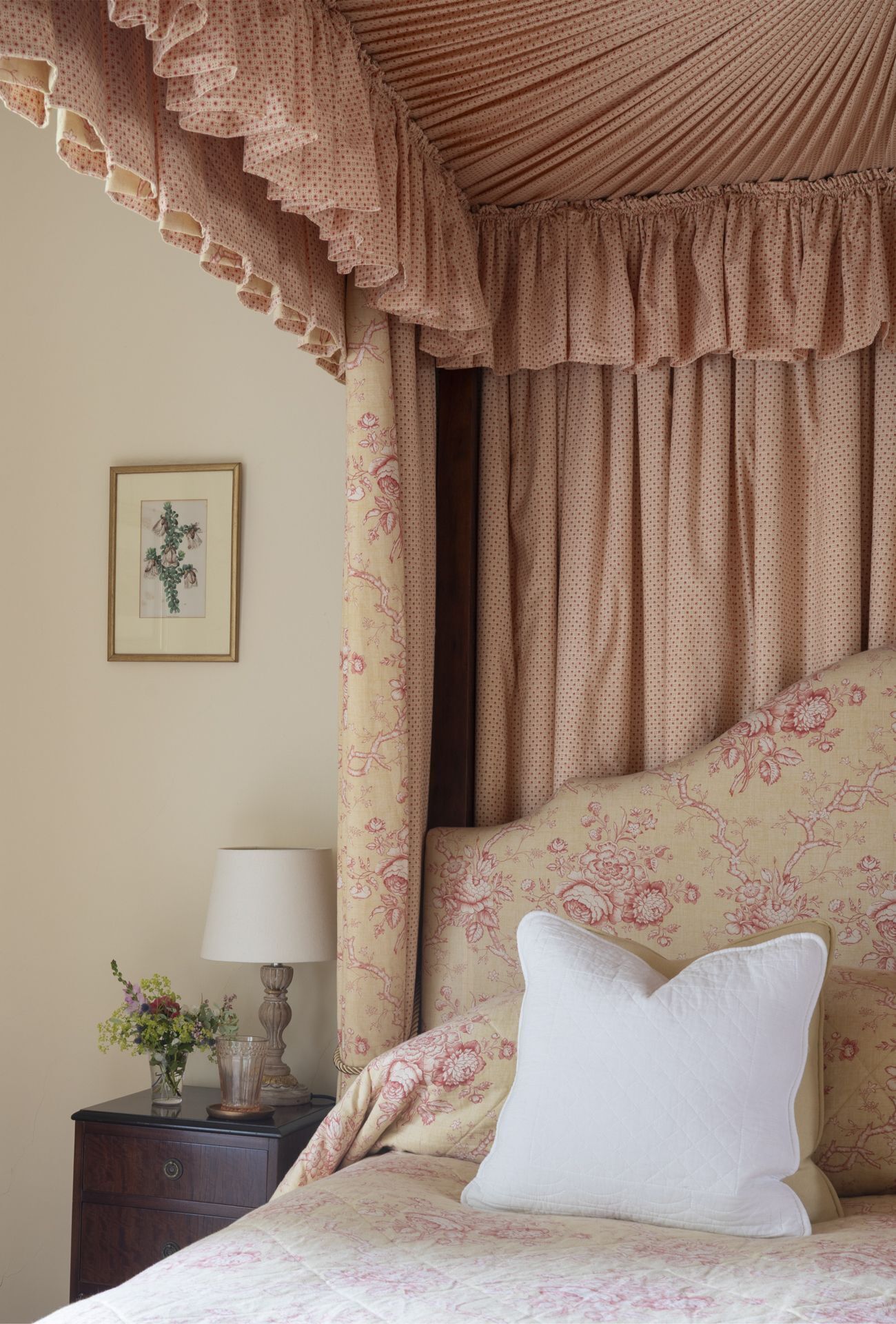 A bedroom with a canopy bed, floral patterned curtains, a bedside table with a lamp, and a framed picture on the wall.