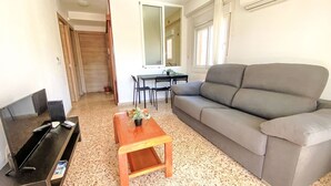 A living room with a grey sofa, a wooden coffee table with a plant, a TV stand, and a dining area with a table and chairs. - Amsterdam Playa Levante by Interhome (Benidorm)