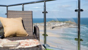 Un balcon avec un fauteuil en osier et un coussin, donnant sur une plage bordée de bâtiments et d’un phare.