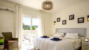 A bedroom with a large bed, a balcony with white curtains, a chair, and a table with a green vase. - Villa Passera by Interhome (Dénia)