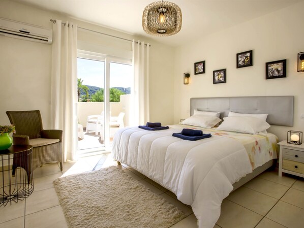 A bedroom with a large bed, a balcony with white curtains, a chair, and a table with a green vase.