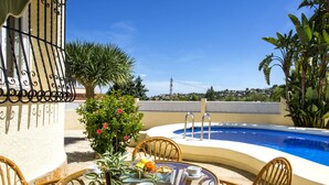 A poolside area with a round table and wicker chairs, surrounded by tropical plants and a clear blue sky. - Villa Passera by Interhome (Dénia)