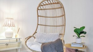A wicker chair with a white cushion, a small round table with a plant and books, and a white lamp on a white console table.