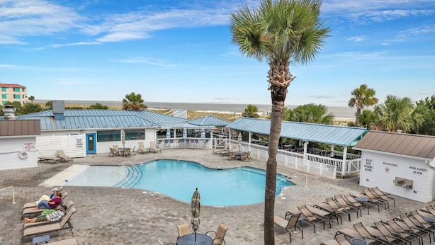 A pool area with lounge chairs, a palm tree, and a building with a blue roof. - Salt & Sea Inn Unit A-1 Bed 1 Ba Condo w/Pool (Tybee Island)