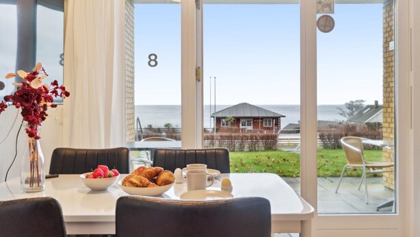 Un coin repas avec une table mise pour le petit-déjeuner, un vase rempli de fleurs et une vue sur la mer, visible à travers de grandes portes vitrées.
