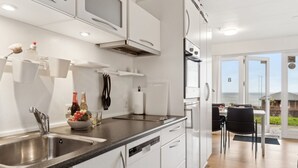Une cuisine moderne avec des meubles blancs, un évier en acier inoxydable et un coin repas offrant une vue sur la mer.