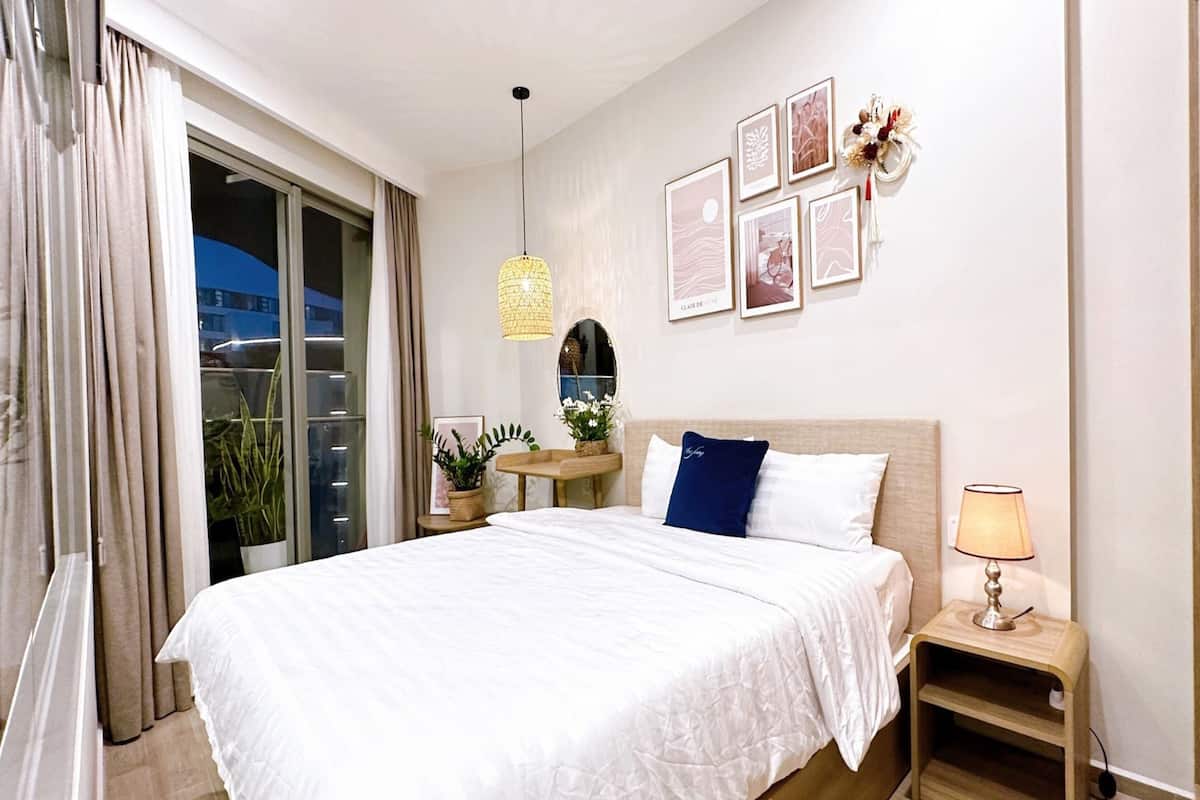 A modern hotel room with a large bed, a bedside table with a lamp, a small side table with a plant, and a wall-mounted mirror.