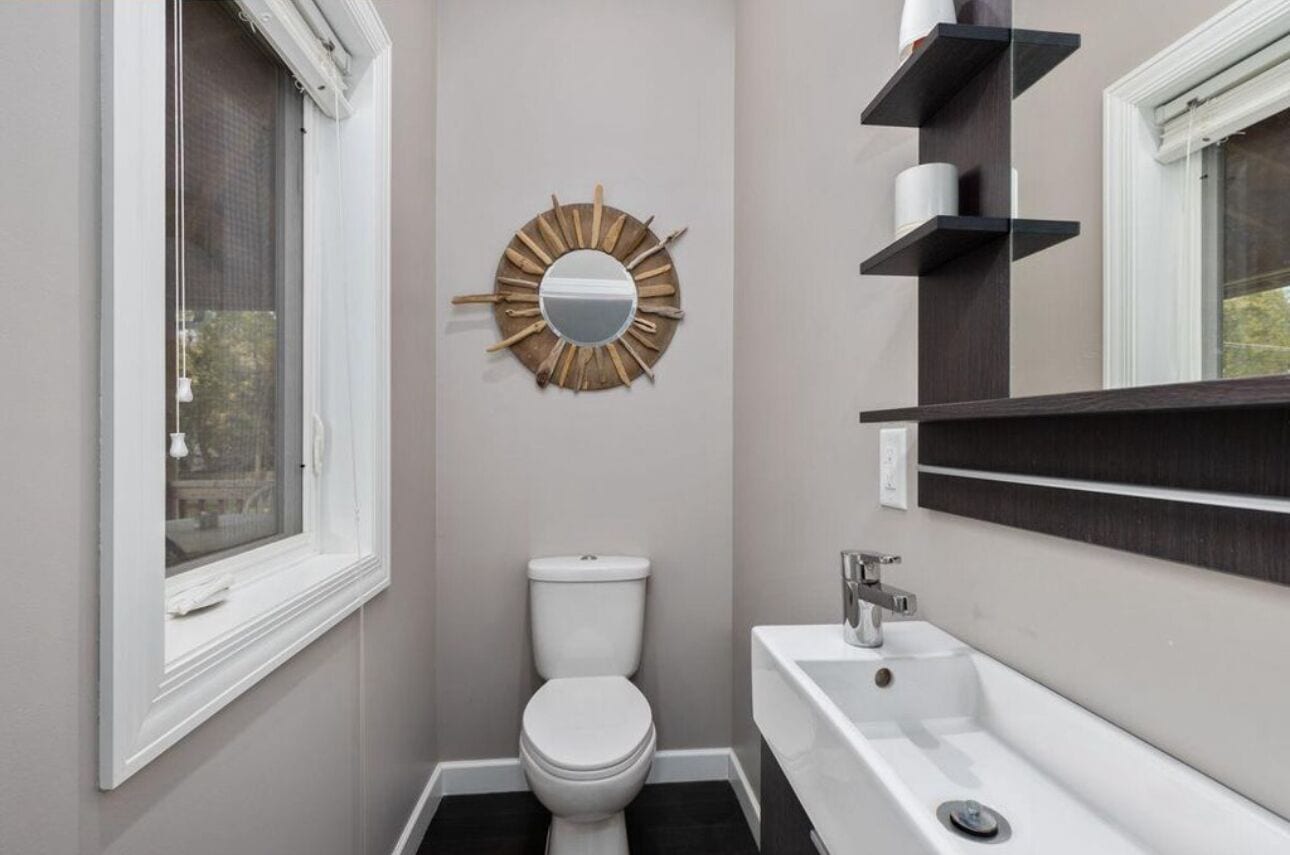 Een kleine badkamer met toilet, een ronde spiegel met houten details en een witte wastafel met donker aanrecht.