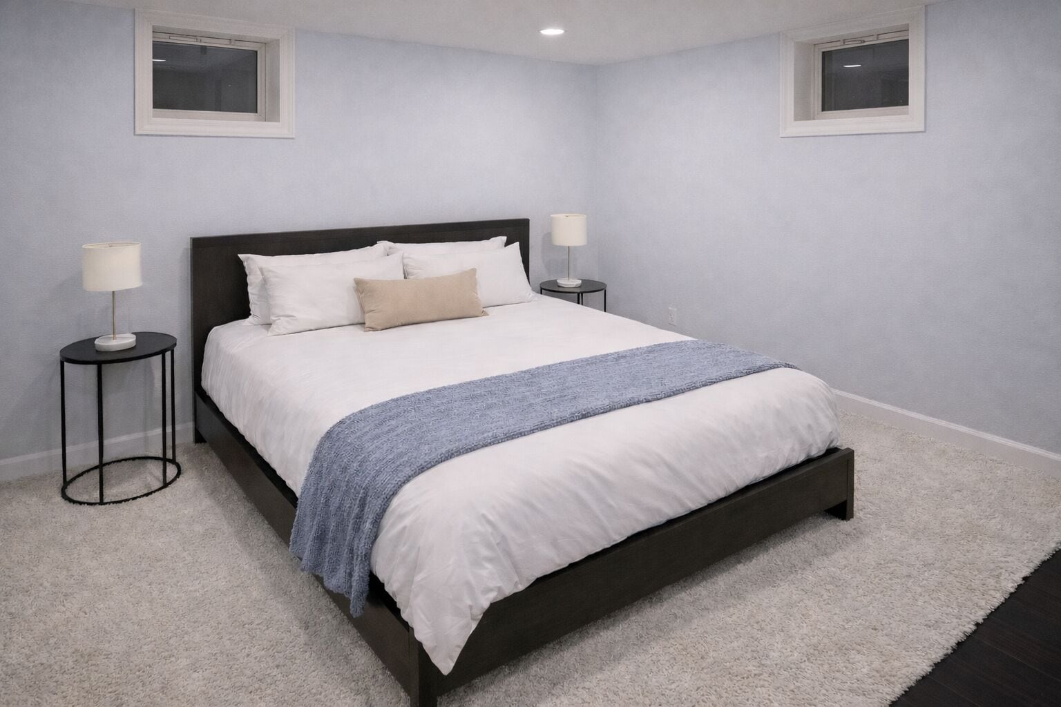 Une chambre avec un lit en bois foncé, du linge de lit blanc, un plaid bleu, deux lampes de chevet et un tapis blanc.