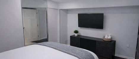 Ett modernt sovrum med en stor säng, ett mörkt konsolbord med en platt-TV och en stor spegel.