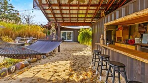 Un espace bar situé en bord de plage, avec un comptoir en bois, des tabourets de bar et un hamac.
