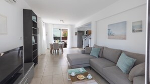 Ein modernes Wohnzimmer mit einer Couch, einem Couchtisch und einem Flachbildfernseher. Im Hintergrund befindet sich ein Essbereich mit Stühlen und einem Tisch.