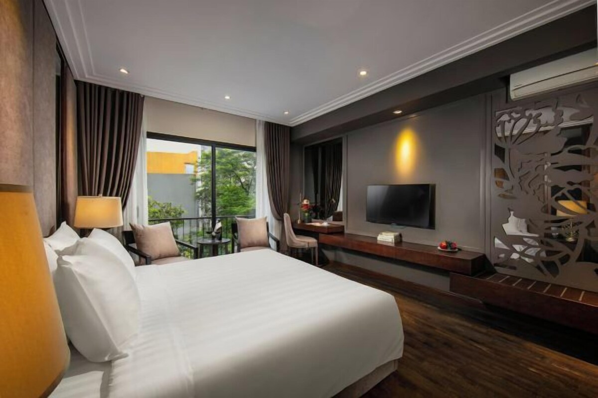 A hotel room with a large bed, a desk with a chair, a TV mounted on the wall, and a balcony with a view of greenery.